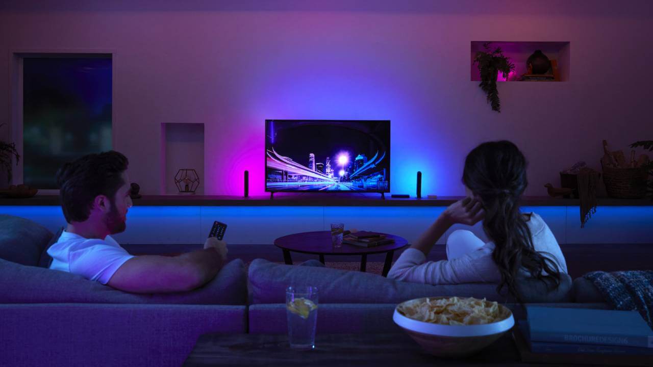 Sepasang pria dan wanita sedang menonton TV dengan lampu-lampu redup dan berwarna
